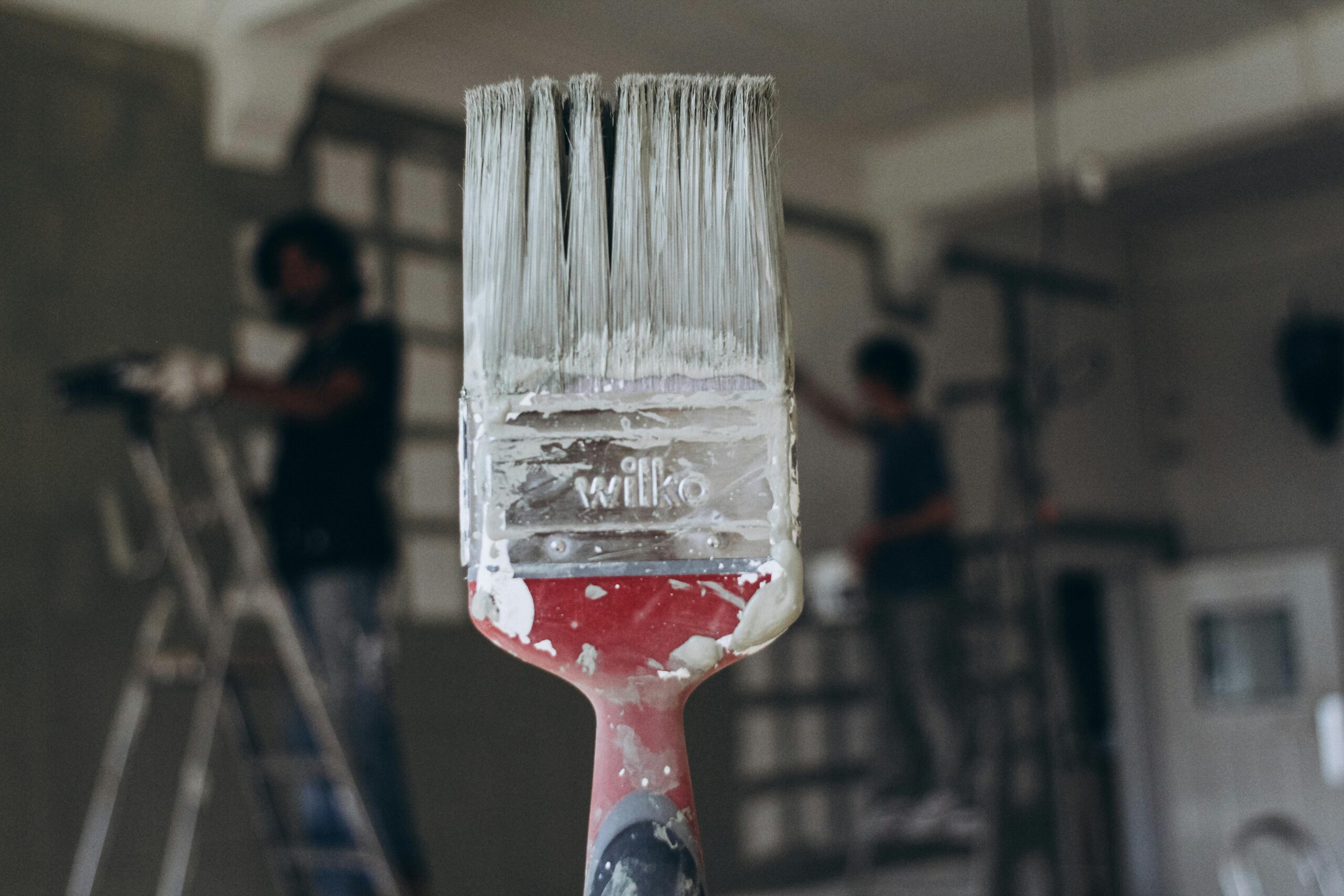 Detailed close-up of a paintbrush with men painting a wall in the blurred background, highlighting renovation work.