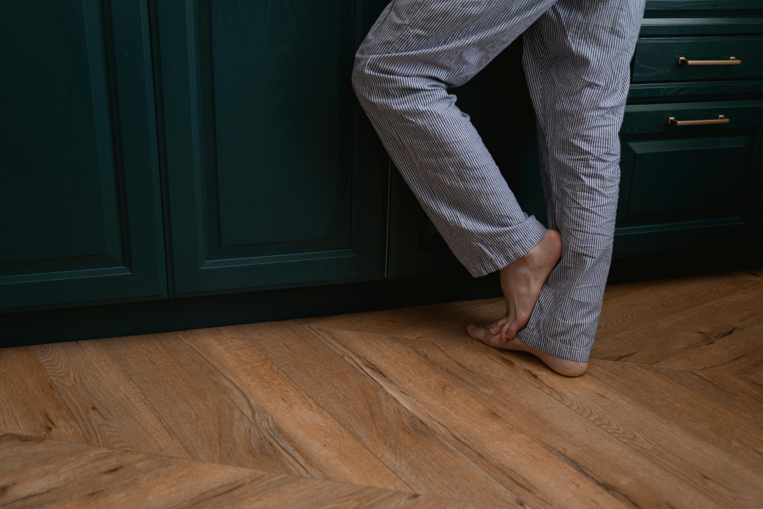Person standing barefoot in striped pajamas on a wooden floor by green cabinets.